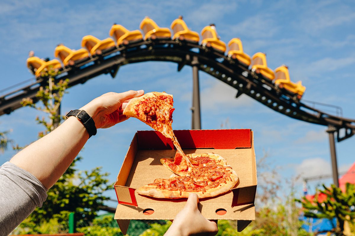 In front of the dino dash ride, a person is holding up a slice of pizza.