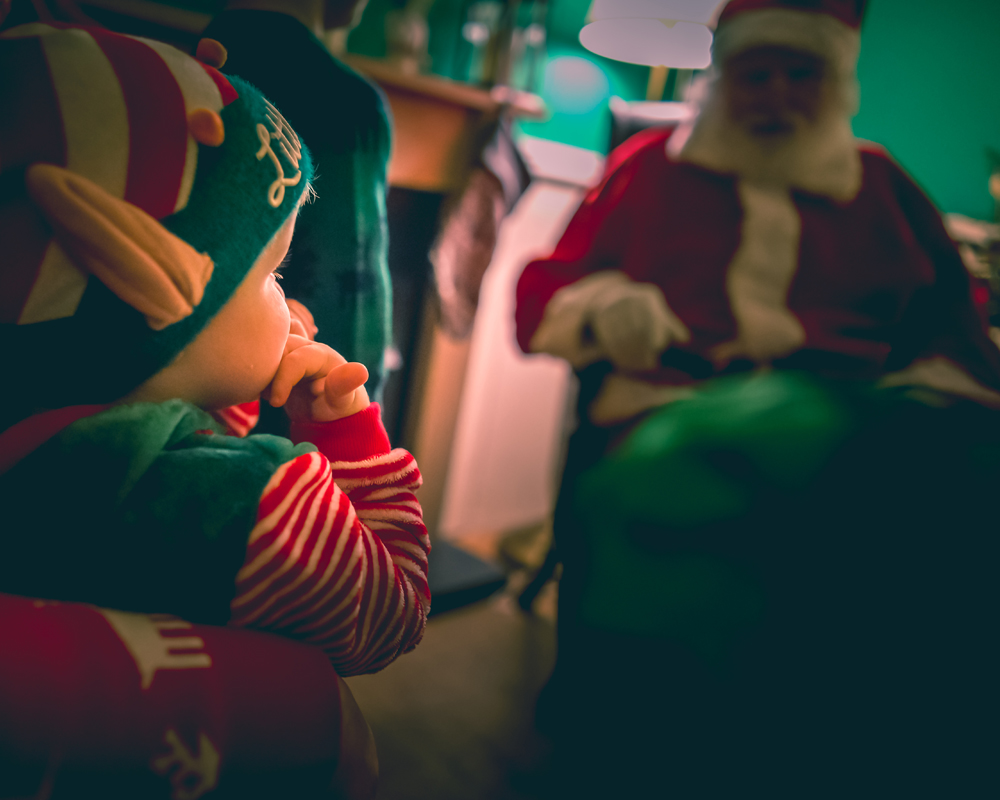 a baby dressed as an elf. Santa Claus is in the background