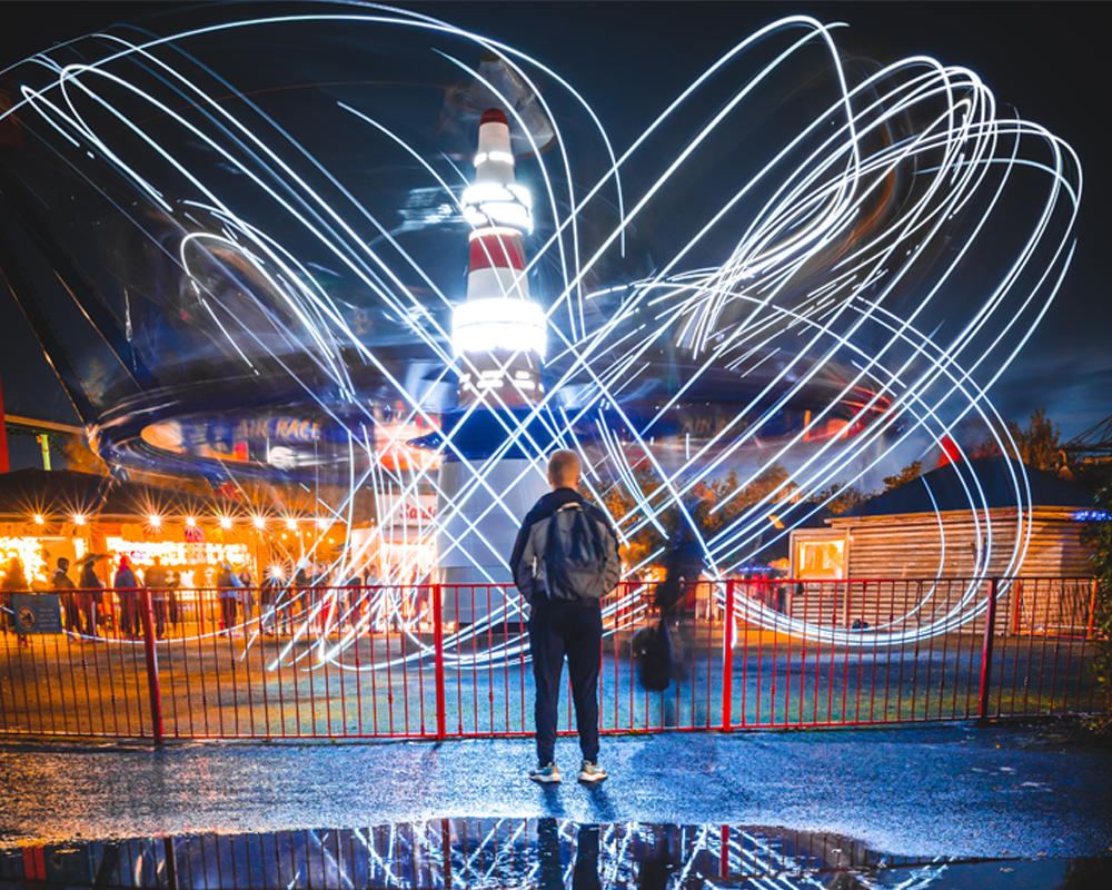 a boy standing with a back pack in front of Air Race at Emerald Park. It is in the dark and the attraction is in motion and lit up.