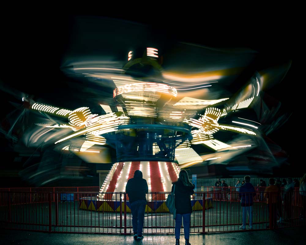 Windstar attraction at Emerald Park. Two guests stand in from of the ride and it is in motion. It is at nighttime and the attraction is lit up.