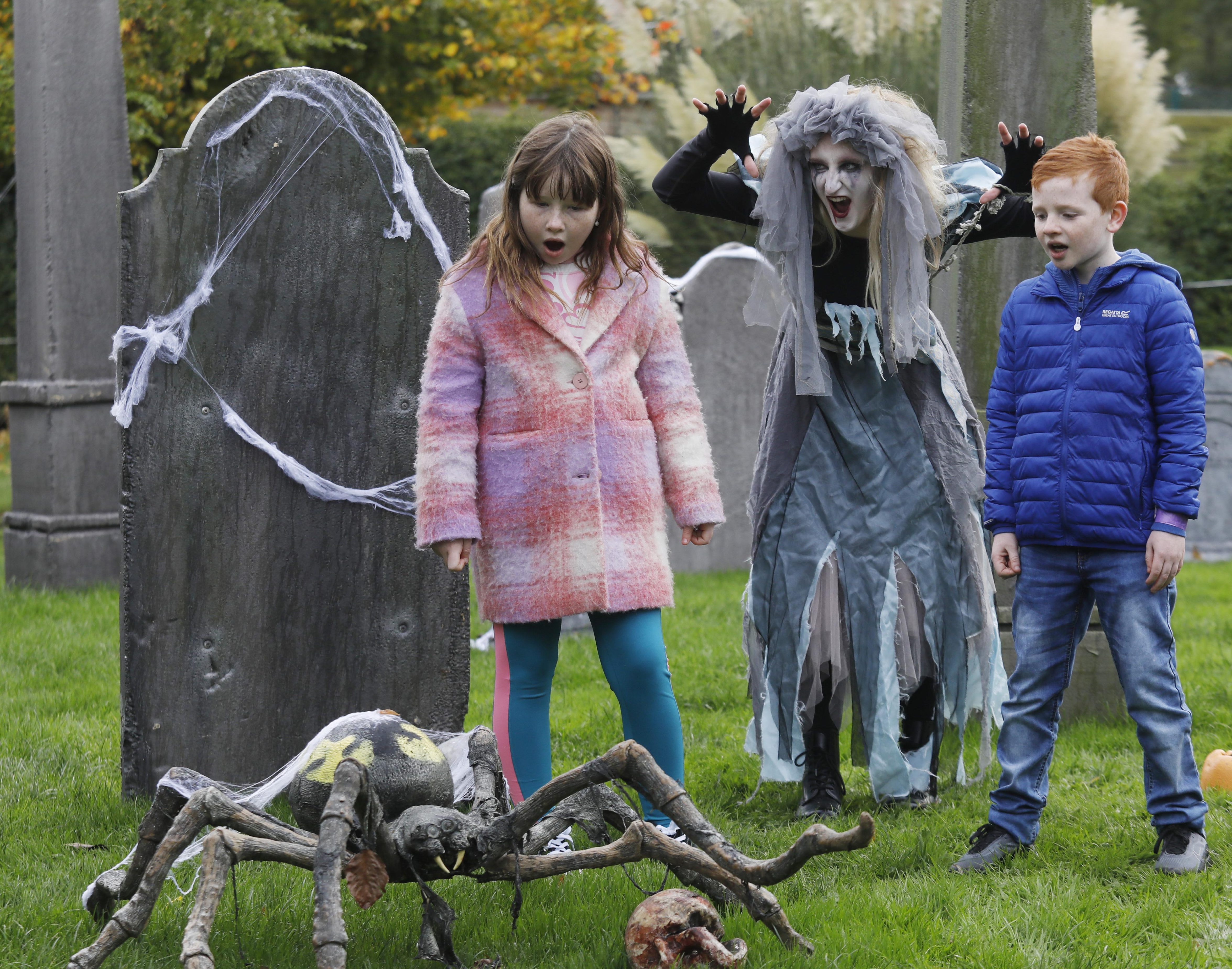 a boy and a girl looking spooked at a spider in a graveyard. A ghoul is behind them