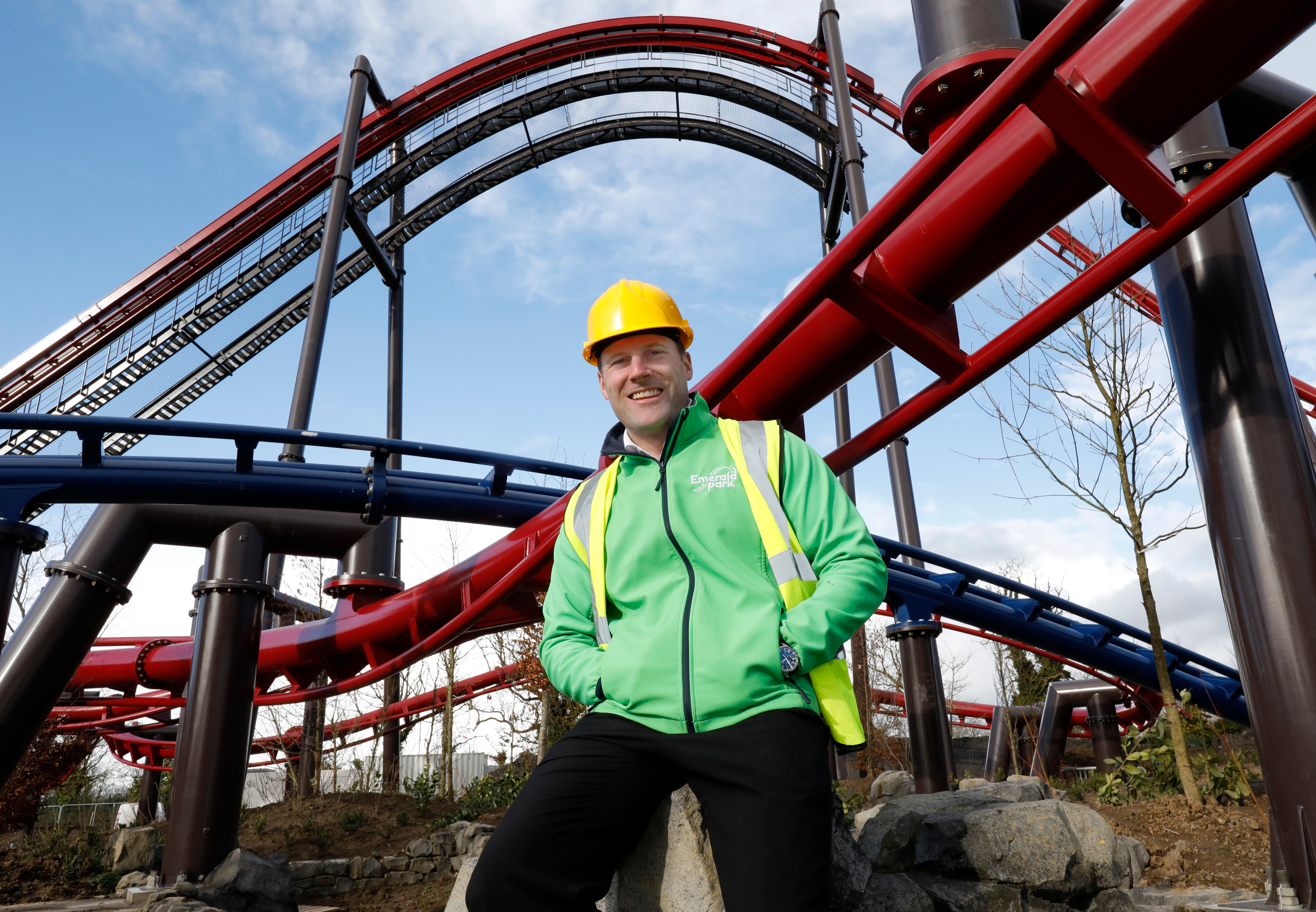 a man wearing a hard hat and an Emerald Park jacket on a boulder with two rollercoasters in the back